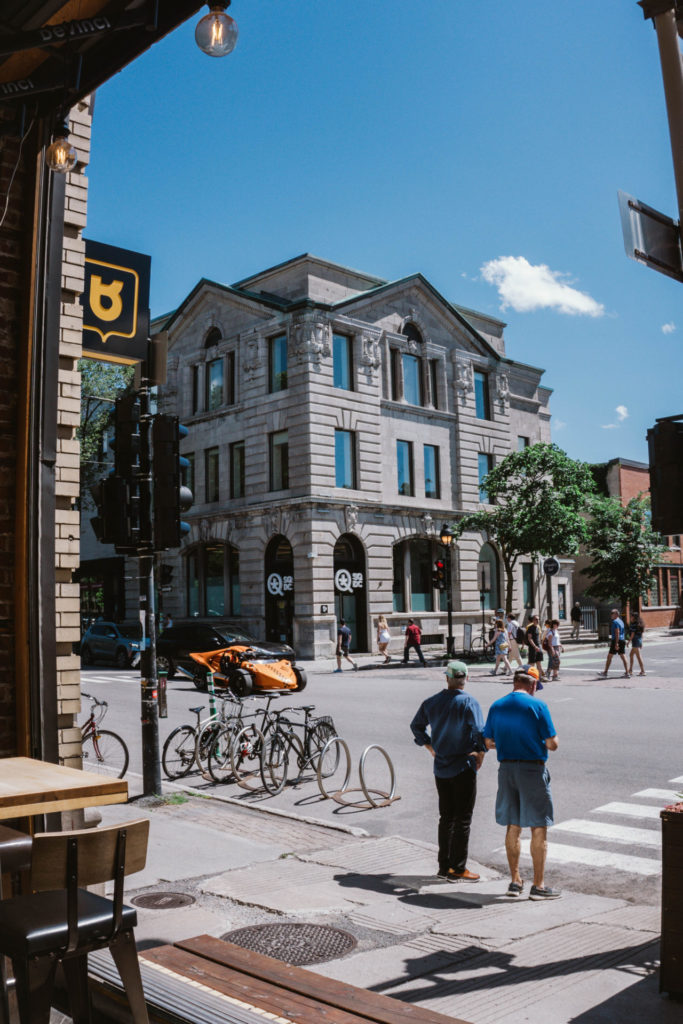 A view of a bustling Montreal street with pedestrians, bicycles, and a striking historic building under a bright blue sky. You can see this view from Siboire Saint Laurent during your Montreal itinerary for 2 days.