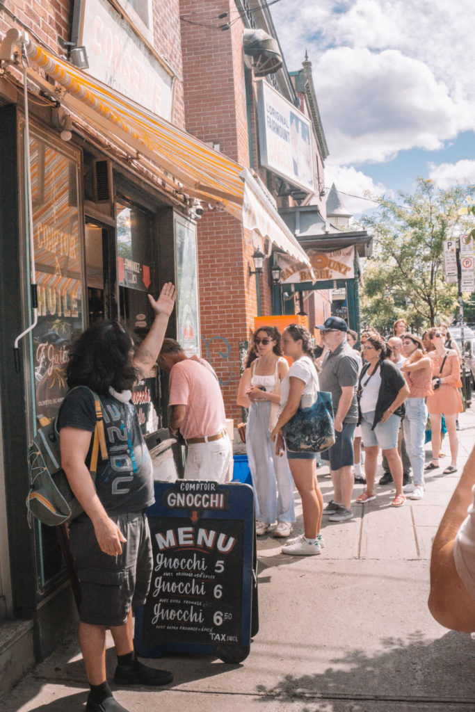 People stand in a line outside Fairmount Bagel Shop, with a "Comptoir Gnocchi" sign and menu board in front. You'll see lots of people queueing up for restaurants during your Montreal itinerary for 2 days.