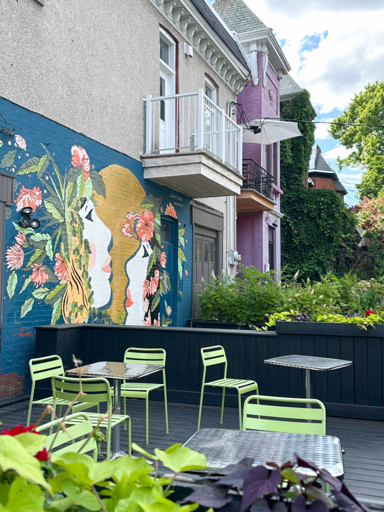 A charming outdoor patio with green metal chairs and tables, adorned with vibrant mural art on the wall depicting floral designs and human profiles. Located in the mile end neighborhood as part of your Montreal itinerary for 2 days.