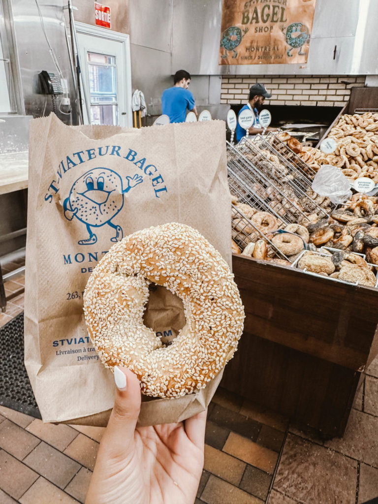 A hand holds a sesame bagel in front of a St-Viateur Bagel paper bag, inside a bagel shop with baskets of fresh bagels and workers in the background. Visiting St-Viateur Bagel is a must for any Montreal itinerary for 2 days.