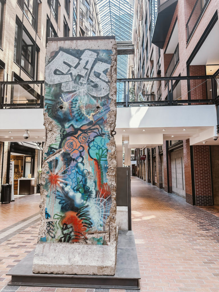 A segment of the Berlin Wall, adorned with graffiti, stands in the atrium of the Montreal World Trade Center, showcasing modern architecture with a glass ceiling and brick walkways. This historical piece makes for an intriguing stop during a quick walkthrough of your Montreal itinerary for 2 days.