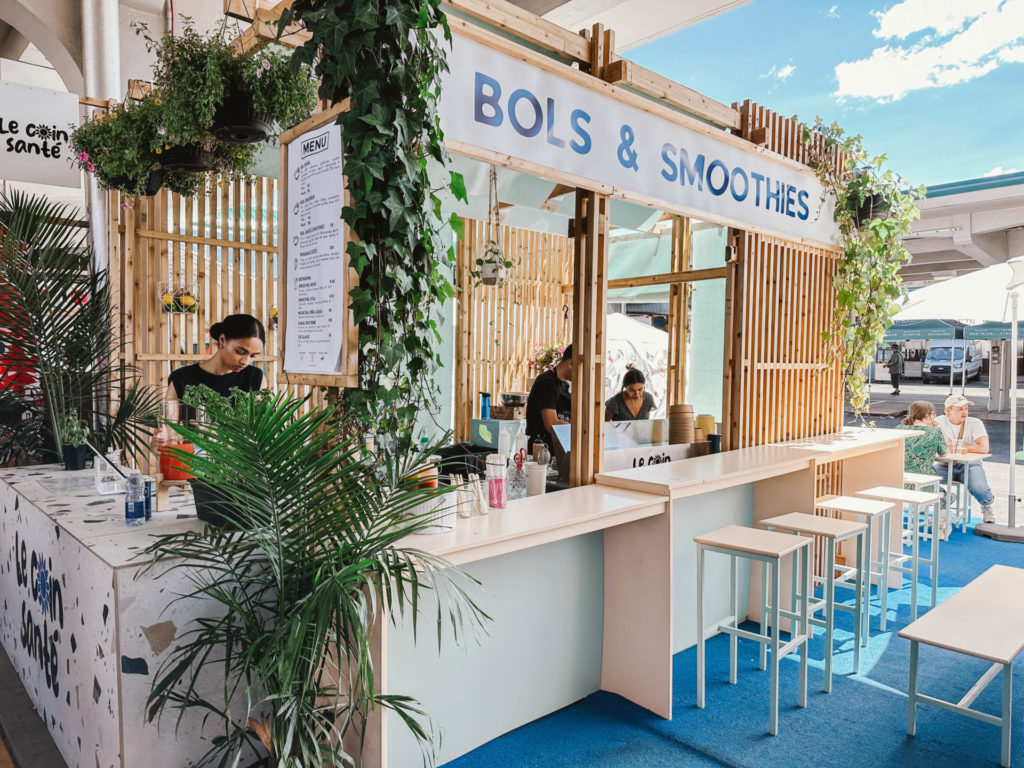 A vibrant smoothie and bowl stand at Jean-Talon Market, with hanging plants and a menu displayed, featuring local vendors and fresh ingredients. Visiting Jean-Talon Market for a taste of local flavors is a great addition to your Montreal itinerary for 2 days.