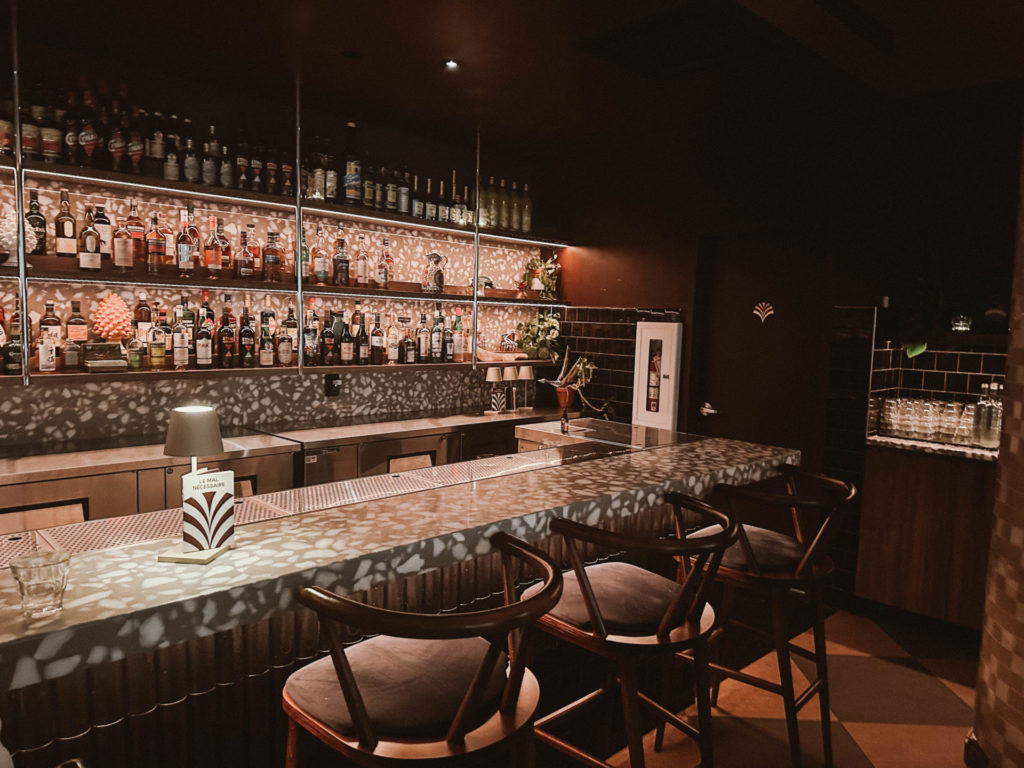 A dimly lit bar with a stylish counter and a wide selection of bottles on illuminated shelves, creating a cozy and inviting atmosphere. Before dinner, treat yourself to cocktails at Le Mal Nécessaire, a perfect addition to your Montreal itinerary for 2 days.