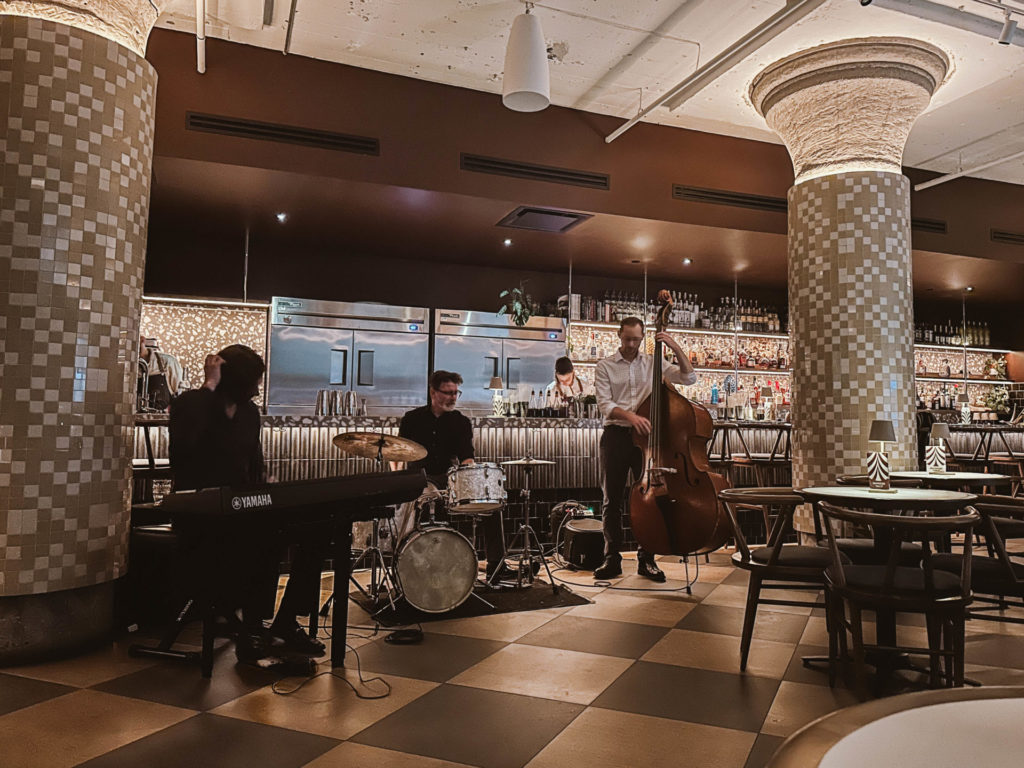 A live band performing at Le Mal Nécessaire, with a pianist, drummer, and double bassist creating a lively ambiance in a stylish bar setting. The live music made the visit 100% worth it, adding to an unforgettable experience in your Montreal itinerary for 2 days.