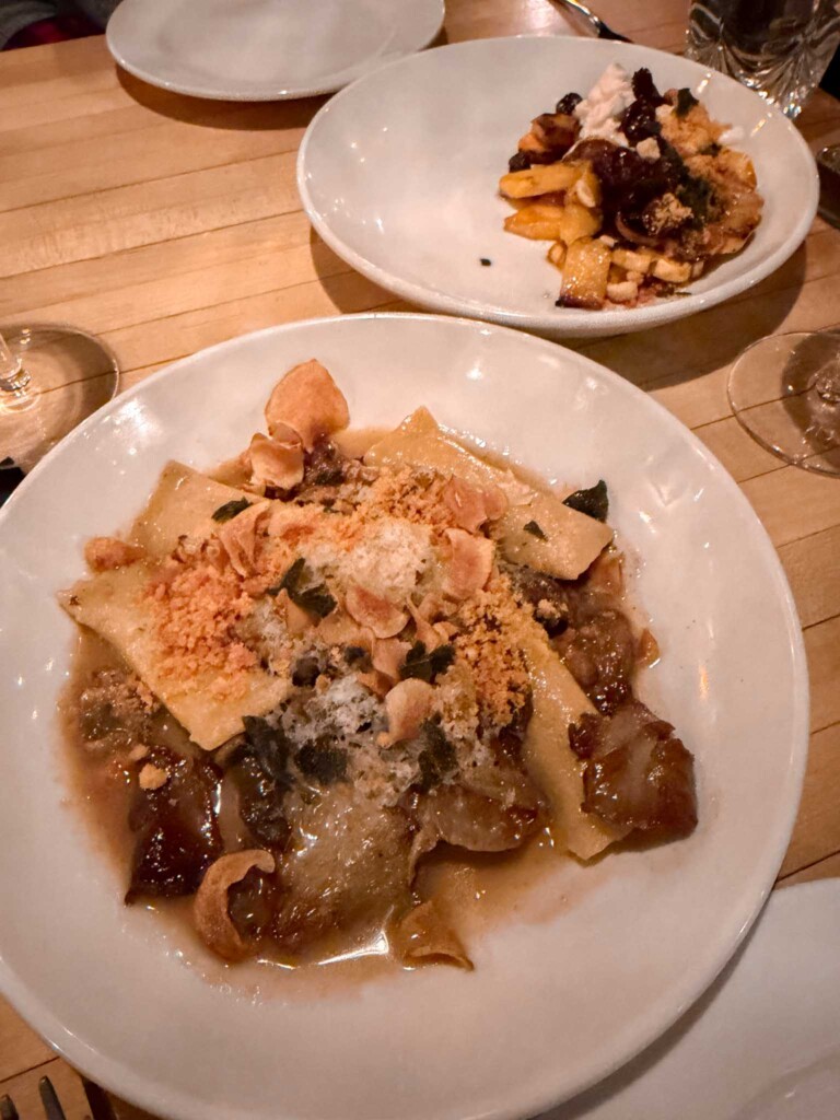 Two white plates on a wooden table feature elegant pasta dishes. The foreground plate holds wide ribbon pasta in a rich brown sauce, topped with shaved cheese, crispy garlic chips, and herbs, while the background plate contains roasted squash with whipped ricotta and a crumble topping.