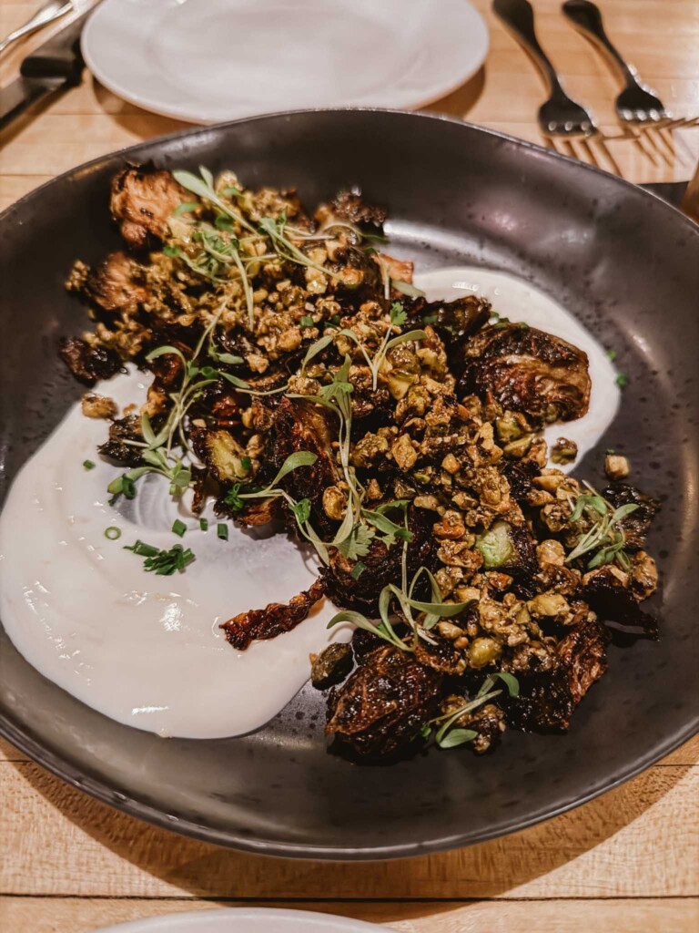 A dark ceramic plate holds a serving of crispy roasted Brussels sprouts topped with chopped nuts and fresh herbs, artfully arranged over a smooth white sauce. The dish is set on a wooden table, with a clean white plate and silverware in the background, suggesting a refined dining experience.