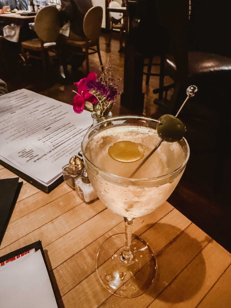 A chilled martini glass sits on a wooden restaurant table, garnished with a lemon twist and a green olive on a cocktail pick. In the background, there's a menu, a salt and pepper shaker set, a small vase with vibrant red and purple flowers, and softly lit dining tables with guests.